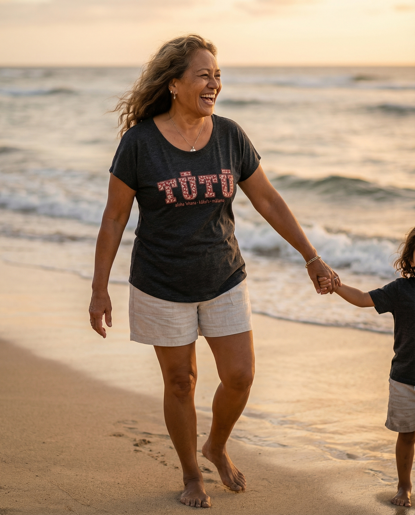 Pua Kukui Tūtū womanʻs dolman shirt in vintage black worn with neutral linen shorts walking on the beach