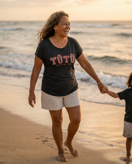 Pua Kukui Tūtū womanʻs dolman shirt in vintage black worn with neutral linen shorts walking on the beach